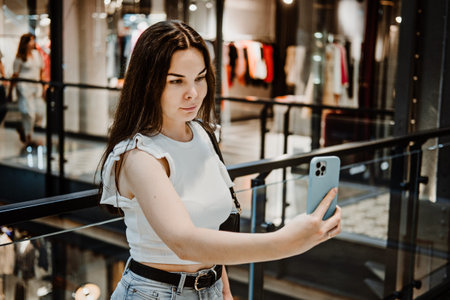 Young Latina woman with shopping bags using her smart phone while standing in shopping center, mall.