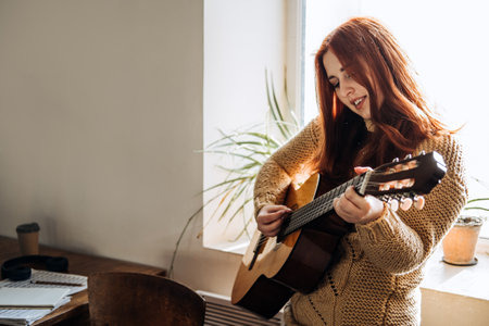 Indoor cozy Hobbies for winter, autumn cold season. Redhead woman playing acoustic guitar and listening to music, sitting near window homeの写真素材