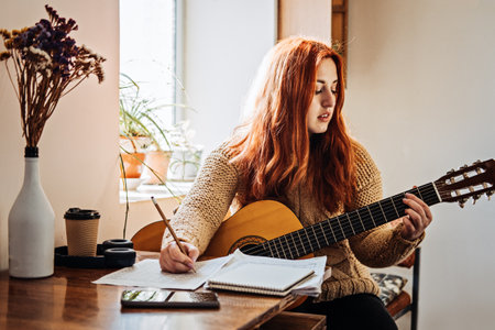 Unaltered candid portrait of young red haired woman in sweater playing acoustic guitar sitting by window at home. Hobbies, indoor home activities for adults in winter, autumn.の写真素材