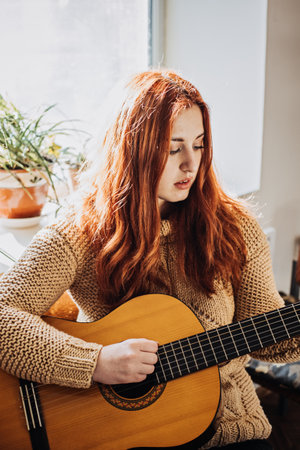 Unaltered candid portrait of young red haired woman in sweater playing acoustic guitar sitting by window at home. Hobbies, indoor home activities for adults in winter, autumn.の写真素材