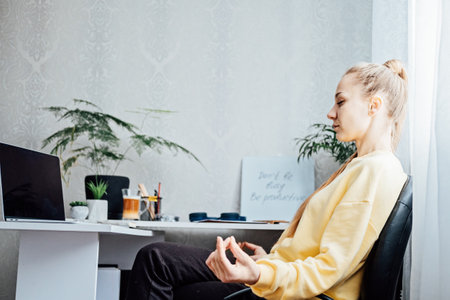 Young woman with close eyes near laptop at home office practicing meditation at table. Freelancer woman meditating near laptop. Taking break time during work. Healing from paperworkの写真素材