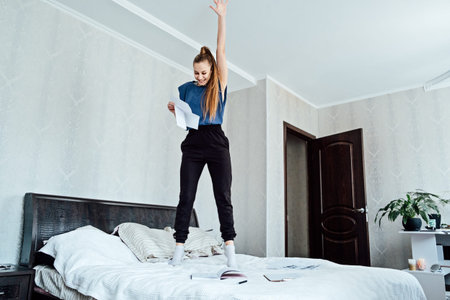 Excited cheerful young woman received a letter, she is hired, jumping on sofa at home, getting good news, feeling joy, dancing, laughing, making winner gesture, happy to win prizeの写真素材