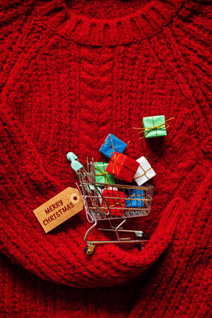 Christmas xmas early sale shopping concept. Shopping cart with many gift boxes on red knitting backgroundの写真素材