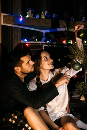 Merry Christmas. Young couple in love celebrating Christmas or new year eve at home. Romantic couple in pajamas sitting and hugging near christmas tree bokeh lights.の写真素材