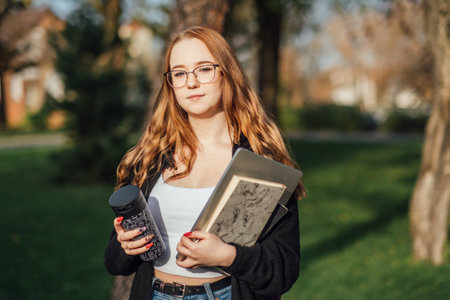 Choosing a university, college. Female college student with books and laptop outdoors. Redhead college student girl with glasses on campusの写真素材