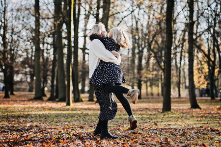 Carefree mother spinning her teenager daughter in autumn day on city public park. Mother and teen daughter playing together in fall parkの写真素材
