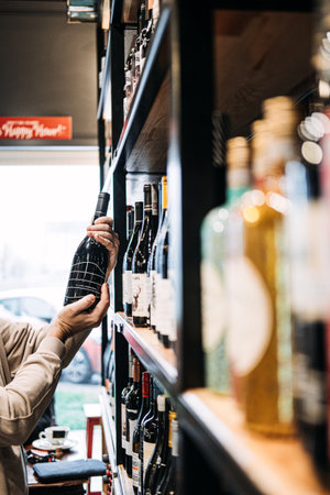 Customer Selecting Wine Bottle from Store Shelf. A persons hand picking a wine bottle from a diverse selection on a well-stocked wine shop shelf. Warsaw, Poland - May 02, 2023.のeditorial素材