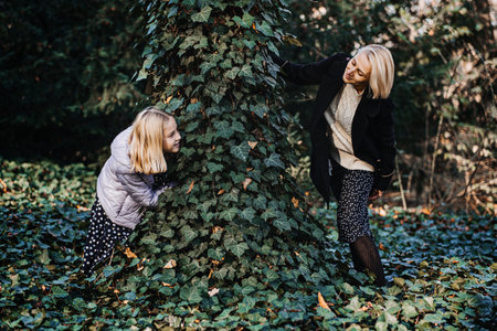 Mother and Daughter Playing Hide and Seek in Park. A joyful mother and daughter engage in a playful game of hide and seek around a tree in an autumn park.の写真素材