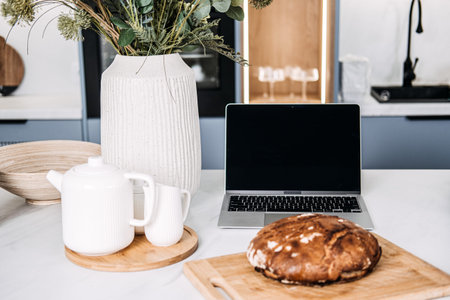 Homemade Bread on Kitchen Table with Laptop. Freshly baked homemade bread on a cutting board beside a laptop, depicting a cozy home baking scene possibly following an online recipe.の写真素材