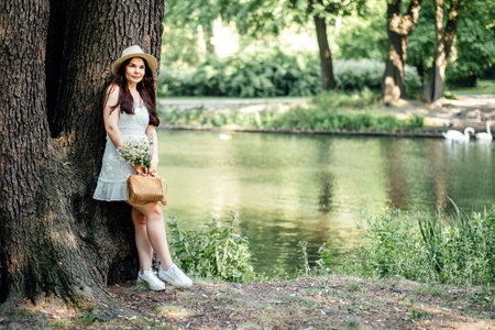 Eco-Friendly Lifestyle. Woman in Nature with Sustainable Fashion Choices. Outdoors woman embodying eco-friendly lifestyle with straw hat, dress, and basket, holding a bouquet of wildflowers.の写真素材