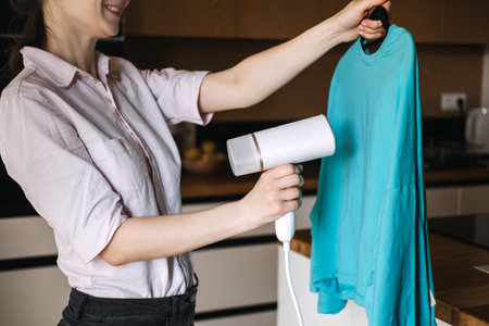 Woman steaming a blue shirt with handheld steamer at home. Garment care, laundry, wrinkle removal, home chores, clothing maintenance.の写真素材