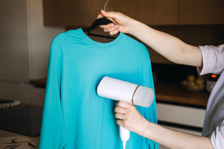 Woman steaming a blue shirt with handheld steamer at home. Garment care, laundry, wrinkle removal,...