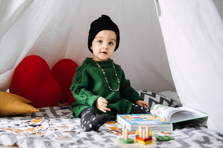 Adorable toddler in green sweater reading book inside a cozy tent. Childhood development, education,...