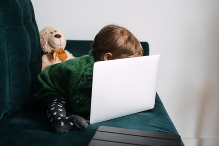 Curious toddler leaning over laptop on couch. Early exposure to technology, childhood curiosity, digital age, learning through play, modern parentingの写真素材