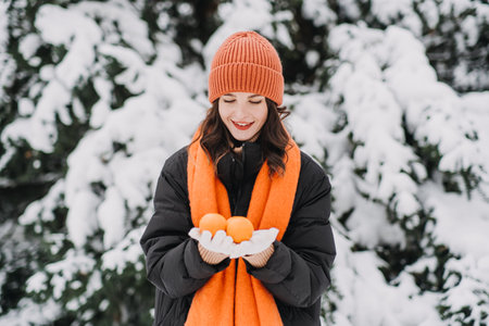 Woman in orange winter attire holding a tangerine, smiling playfully in snow-covered surroundings. Winter wellness, mindfulness, and seasonal joy.の写真素材