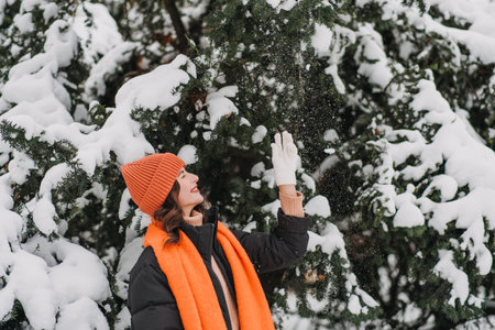 Young woman in orange winter accessories reaching for snow on tree branches. Forest bathing, mindful winter walks, outdoor self-care trendsの写真素材