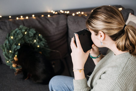 Woman taking photo of black cat hiding in Christmas wreath with string lights on couch. Cozy indoor...