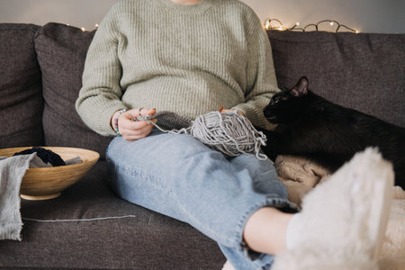 Woman knitting with black cat on sofa, embracing cozy downtime. Relaxation, companionship, digital detox, and mindful hobbies concept.の写真素材