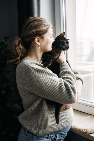 Pregnant woman holding black cat by the window, questioning the safety of keeping pets during pregnancy. Cozy home with coffee and candle.の写真素材