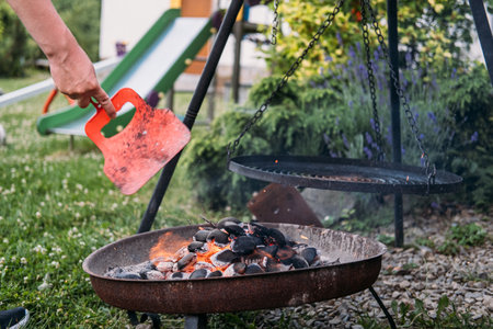 Outdoor fire pit with glowing coals and a hand using a red fan to stoke the flames, surrounded by a backyard settingの写真素材