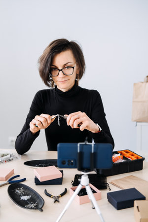 Handmade jewelry business owner recording a video tutorial with a smartphone. Content creation, social media marketing, small business, digital entrepreneurship, online branding.の写真素材