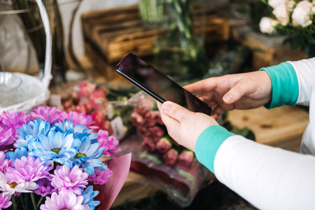 Florist using smartphone in flower shop to manage digital storefront. Reflects ecommerce floristry, small business tech, and mobile marketing trendsの写真素材