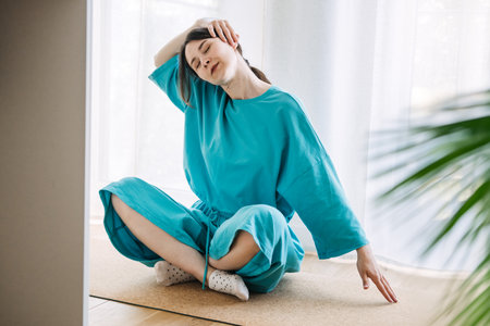 Young white woman in a turquoise robe does a neck stretch while sitting cross-legged on a mat in a bright room. Soft wellness, mindful morning routine, home self-care, body awarenessの写真素材