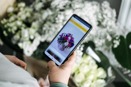 Person browsing a flower bouquet on a smartphone screen surrounded by real white blossoms. Flower subscription, floral e-commerce, gift delivery services, digital floristryの写真素材