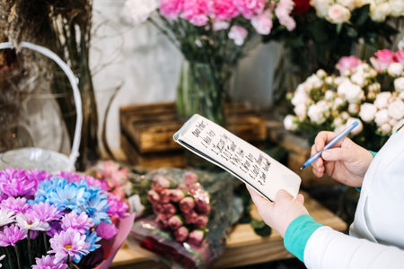 Florist writing down open bouquet orders in a notepad while surrounded by vibrant floral arrangements. Small business workflow, order management, floral studio, creative entrepreneurの写真素材