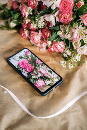Smartphone displaying a bouquet photo next to real pink flowers on a kraft paper background. E-commerce floristry, digital product showcase, florist branding, online flower businessの写真素材
