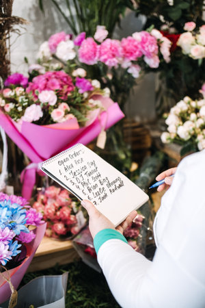 Florist writing down open bouquet orders in a notepad while surrounded by vibrant floral arrangements. Small business workflow, order management, floral studio, creative entrepreneurの写真素材