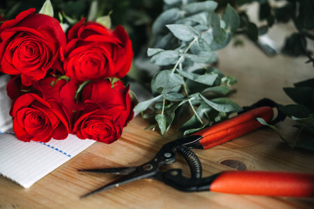 Red roses, eucalyptus, and pruning shears arranged on a florists wooden worktable. Floral arranging, hands-on creativity, rose bouquet, artisan florist, working process. High quality photoの写真素材