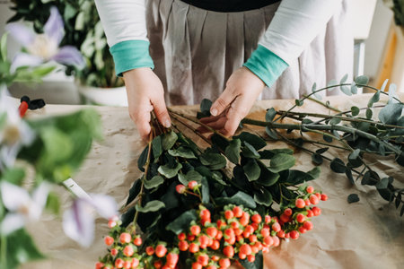 Florist creating a bouquet with orange berries and eucalyptus branches on a kraft-covered table....