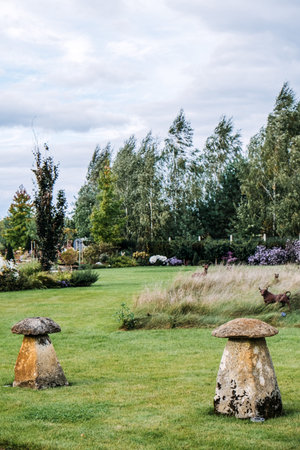 Two large mushroom-shaped stone sculptures placed on a manicured lawn within a landscaped garden featuring wild grasses and floral borders. Garden ornamentation, natural landscapingの写真素材