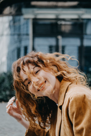 A young woman with curly red hair smiles joyfully as the wind tosses her hair in front of her face....
