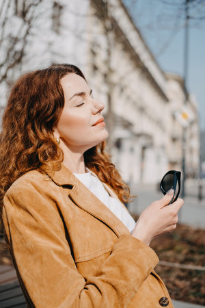 Young woman with auburn curly hair peacefully basking in the sun on a city sidewalk. City life wellness, urban pause, mental reset, mindful moments...の写真素材