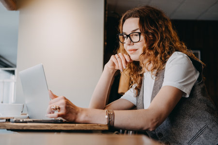 A young woman with curly red hair concentrates on her laptop in a softly lit cafe. Focused productivity, deep work sessions, digital workflow, cognitive engagement...の写真素材