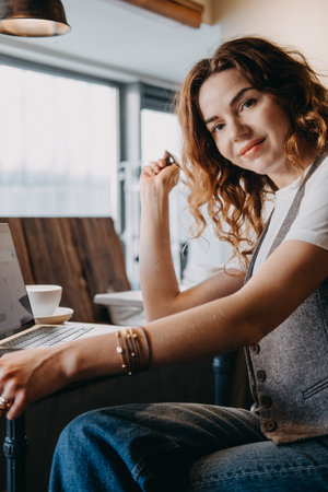 A young woman with curly red hair pauses work to smile at the camera in a cozy cafe setting. Women in digital professions, female freelancer, tech industry woman, creative remote worker...の写真素材