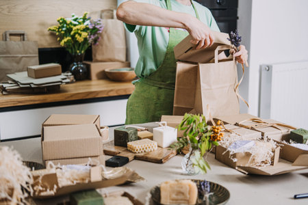 Gift boxes with handmade soaps are prepared on a kitchen table while a person ties a kraft bag. DIY...