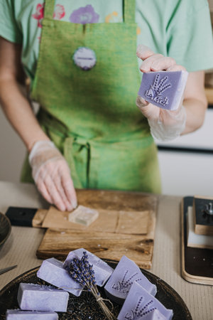 Person in green apron stamps a lavender soap bar by hand on a wooden board. Handmade soap branding, artisan process, small batch skincare, craft identityの写真素材