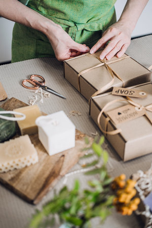 Person ties ribbon on kraft soap boxes beside artisanal bars and eco materials. Zero waste...