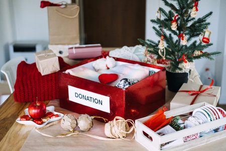 DONATION box filled with clothes stands on table beside Christmas tree and wrapped gifts. Christmas donation box, holiday charity clothing drive, donating warm clothes winter.