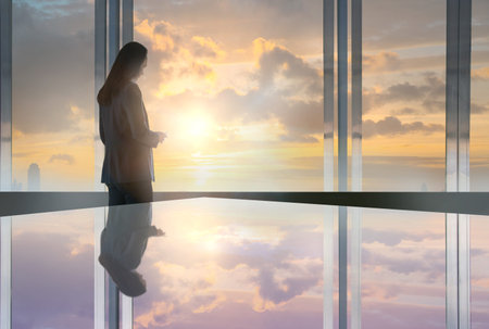 Young woman standing next to the office window and reading message on the phoneの写真素材