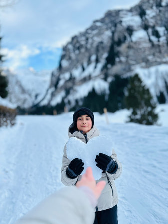Cute little boy in winter clothes playing with snow in the mountains. A little boy holds a heart made of snow against the backdrop of the Swiss snowy mountainsの写真素材