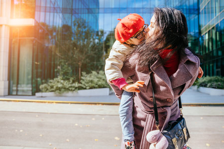 Cute young daughter on a piggy back ride with her mother in autumn time with modern building at city street on background. Sun glare efectの写真素材