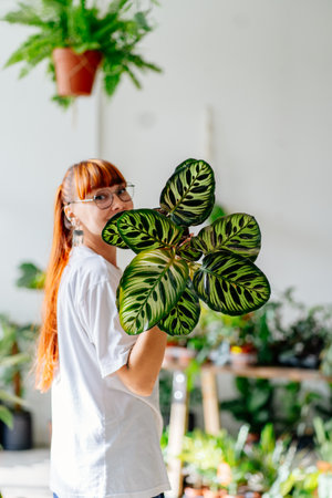 Sustainable lifestyle, slow living concept. Young woman with red hair florist, plant lover, hide her face with plant in pot in greenery floral store boutique.の写真素材