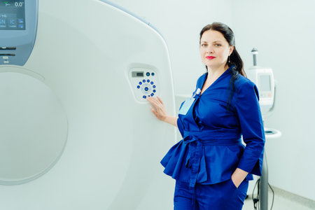 A woman stands in front of a GE MRI machine. She is wearing a blue uniform and she is a medical professionalの写真素材