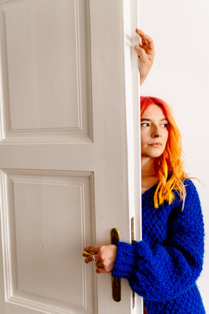 Creative woman with orange hair stands in front of white door. She is wearing a blue sweater and has a ring on her finger. The door is open, and the woman is looking out. Vertical.の写真素材