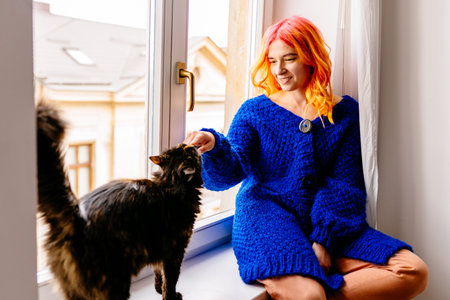 A woman with orange hair is petting a cat while wearing a blue cozy sweater. The cat is sitting on a windowsill. Pet lover concept.の写真素材