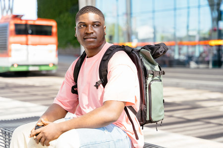 African student man traveler wearing a pink shirt and a backpack sits on a bench waiting for bus...